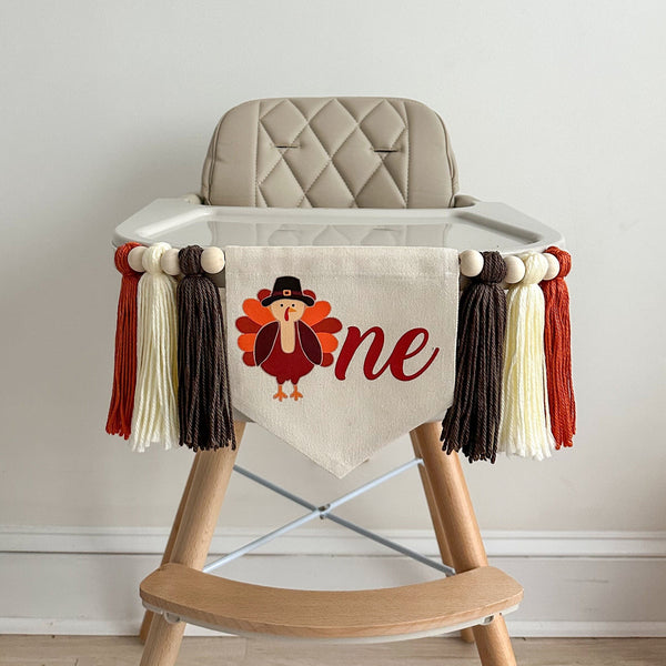 A 5 by 7 inch canvas center flag with a fun turkey followed by the letters NE in deep red to make the word ONE. There is a taupe brown, cream and dark orange 5 inch yarn tassel on each side. Each tassel is separated by a wooden bead.