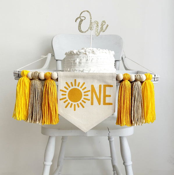 A 5 by 7 inch canvas flag with a sunshine followed by the letters NE in yellow. There is a 5 inch gold, tan and yellow yarn tassel on each side, each separated by a wooden bead for a total of 6 tassels and 6 beads.