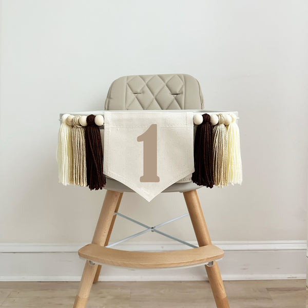 A 5 x 7 inch canvas flag with a tan number 1 on it. On each side is a brown, tan and cream colored 5 inch yarn tassel each separated by a wooden bead. There is a total of 6 tassels and 6 wooden beads.