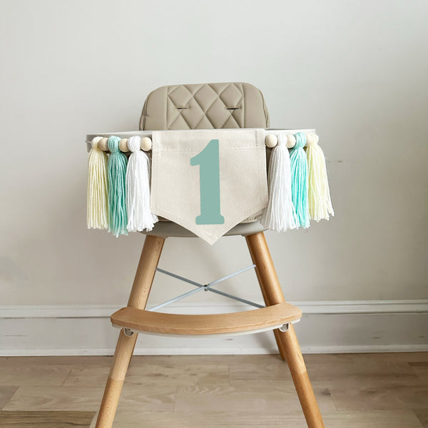 A 5 x 7 inch canvas flag with a mint green number 1 on it. On each side is a white, mint green and cream colored 5 inch yarn tassel each separated by a wooden bead. There is a total of 6 tassels and 6 wooden beads.
