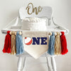 A 5 by 7 inch canvas center flag with a red and white fishing bobber followed by the letters NE in navy blue to spell the word ONE. On each side is a 5 inch light blue, tan and red yarn tassel each separated by a wooden bead for a total of 6 tassels.