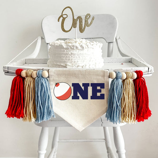 A 5 by 7 inch canvas center flag with a red and white fishing bobber followed by the letters NE in navy blue to spell the word ONE. On each side is a 5 inch light blue, tan and red yarn tassel each separated by a wooden bead for a total of 6 tassels.
