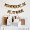 A burlap banner made from 4.5 by 6.5 inch fishtail shaped flags. The word ROOKIE is on the top line in black followed by a baseball image and OF, and the bottom line has the word THE followed by a baseball image and the word YEAR in black.
