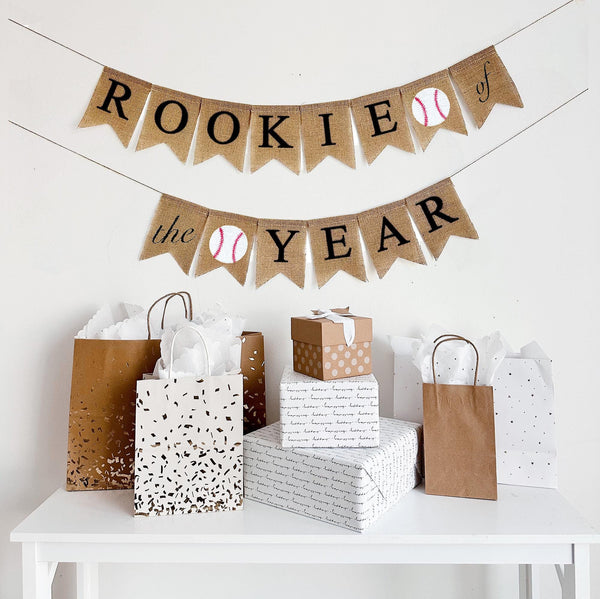 A burlap banner made from 4.5 by 6.5 inch fishtail shaped flags. The word ROOKIE is on the top line in black followed by a baseball image and OF, and the bottom line has the word THE followed by a baseball image and the word YEAR in black.