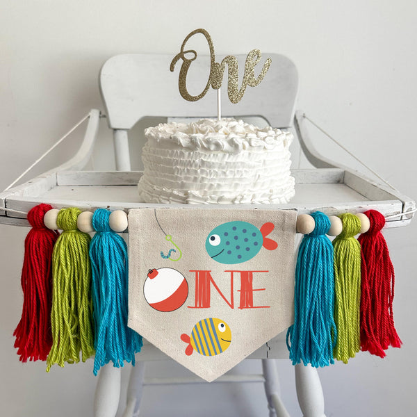 A 5 by 7inch canvas center flag with a fishing bobber and the letters NE to spell ONE with a blue fish and a yellow tropical style fish as accents. There is an aqua, lime and red 5 inch yarn tassel on each side, each separated by a wooden bead.