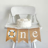 A burlap banner made from three 5 by 7 inch fishtail shaped flags. The first flag has a white buoy lifesaver with navy blue stripes. The second flag has a white lowercase N. The third flag has a white lowercase E. It is shown on a high chair.