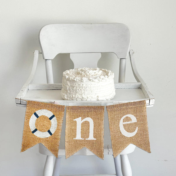 A burlap banner made from three 5 by 7 inch fishtail shaped flags. The first flag has a white buoy lifesaver with navy blue stripes. The second flag has a white lowercase N. The third flag has a white lowercase E. It is shown on a high chair.