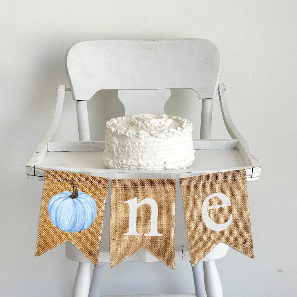 A burlap banner made from three 5 x 7 inch fishtail shaped flags. The first flag has a blue watercolor style pumpkin on it to create the letter O. The second flag has a lowercase letter N in white and the third flag has a lowercase letter E in white.