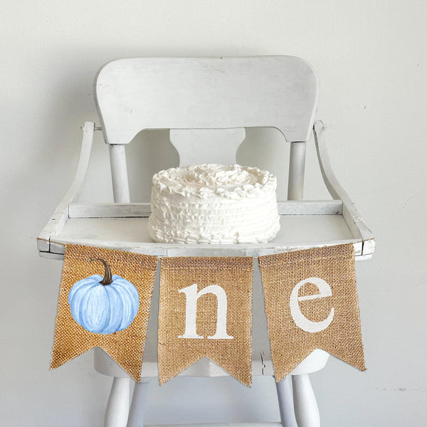 A burlap banner made from three 5 x 7 inch fishtail shaped flags. The first flag has a blue watercolor style pumpkin on it to create the letter O. The second flag has a lowercase letter N in white and the third flag has a lowercase letter E in white.