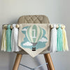 A 5 by 7 inch canvas center flag with a mint and creamy yellow striped hot air balloon and clouds. There is a 5 inch white, mint green and cream colored yarn tassel on each side each separated by a wooden bead for a total of 6 tassels.