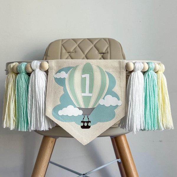 A 5 by 7 inch canvas center flag with a mint and creamy yellow striped hot air balloon and clouds. There is a 5 inch white, mint green and cream colored yarn tassel on each side each separated by a wooden bead for a total of 6 tassels.