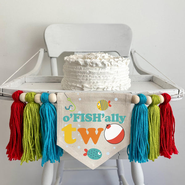 A 5 by 7 inch canvas center flag with the words OFISHALLY TWO with a bobber as the O and 2 tropical fish and bubbles. There is a 5 inch aqua, lime and red colored yarn tassel on each side each separated by a wooden bead for a total of 6 tassels.
