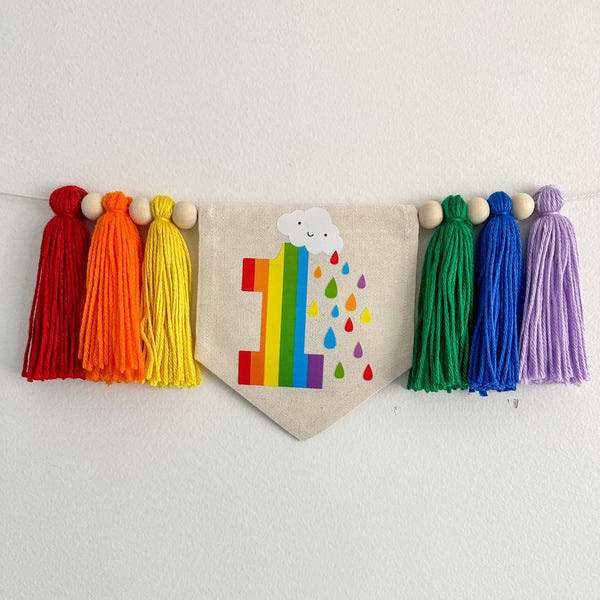 A 5 by 7 inch canvas center flag with a rainbow number 1, a cloud and rain drops. There is a 5 inch red, orange, yellow, green, blue and lavender colored yarn tassel on each side each separated by a wooden bead for a total of 6 tassels.