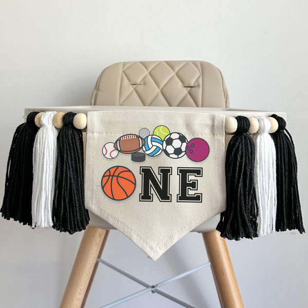 A 5 by 7 inch canvas center flag with a basketball and the letters NE in black with a row of sports balls at the top. There is a 5 inch black, white and black colored yarn tassel on each side each separated by a wooden bead for a total of 6 tassels.