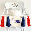 A 5 by 7 inch canvas banner with a baseball as the O and the letters NE in navy to spell ONE. There is a 5 inch navy, white and red yarn tassel on each side with a wooden bead between each of them for a total of 6 tassels.
