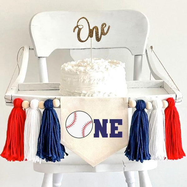 A 5 by 7 inch canvas banner with a baseball as the O and the letters NE in navy to spell ONE. There is a 5 inch navy, white and red yarn tassel on each side with a wooden bead between each of them for a total of 6 tassels.