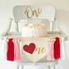 A 5 by 7 inch canvas center flag with a red glitter heart as the O and the letters NE in gold glitter to spell ONE. There is a 5 inch pink, red and white yarn tassel on each side, each separated by a wooden bead.
