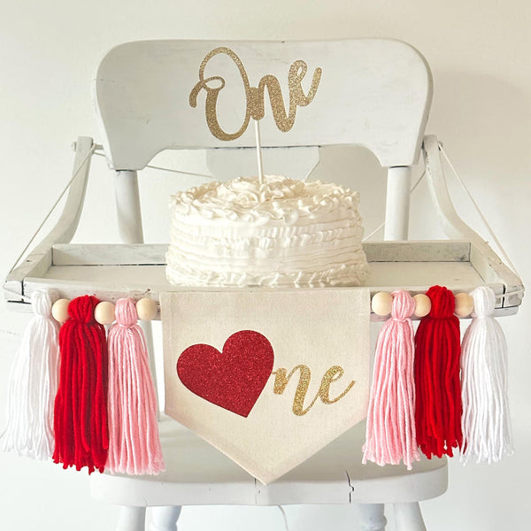 A 5 by 7 inch canvas center flag with a red glitter heart as the O and the letters NE in gold glitter to spell ONE. There is a 5 inch pink, red and white yarn tassel on each side, each separated by a wooden bead.