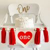 A 5 by 7 inch center flag with a red heart and the word ONE inside the heart. There is a red, white and red 5 inch yarn tassel on each side, each separated by a wooden bead for a total of 6 tassels. It is shown on a white wooden high chair.
