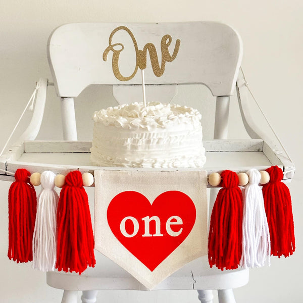A 5 by 7 inch center flag with a red heart and the word ONE inside the heart. There is a red, white and red 5 inch yarn tassel on each side, each separated by a wooden bead for a total of 6 tassels. It is shown on a white wooden high chair.