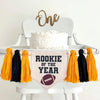 A 5 by 7 inch canvas center flag with the words ROOKIE OF THE YEAR in black with a football at the bottom. There is a 5 inch gold, black and gold yarn tassel on each side each separated by a wooden bead for a total of 6 tassels.