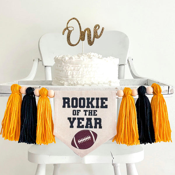 A 5 by 7 inch canvas center flag with the words ROOKIE OF THE YEAR in black with a football at the bottom. There is a 5 inch gold, black and gold yarn tassel on each side each separated by a wooden bead for a total of 6 tassels.