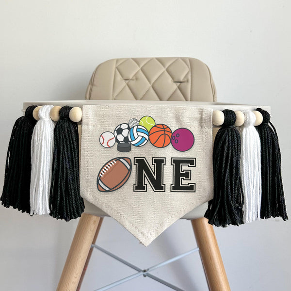 A 5 by 7 inch canvas center flag with a football and the letters NE in black with a row of sports balls at the top. There is a 5 inch black, white and black colored yarn tassel on each side each separated by a wooden bead for a total of 6 tassels.