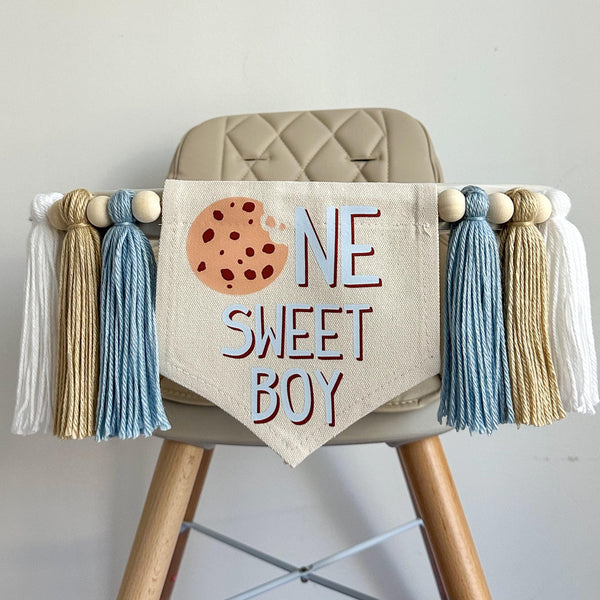 A 5 by 7 center flag with a chocolate chip cookie followed by the letters NE in light blue and SWEET BOY in light blue. On each side there are 5 inch light blue, tan and white tassels each separated by a wooden bead for a total of 6 tassels.