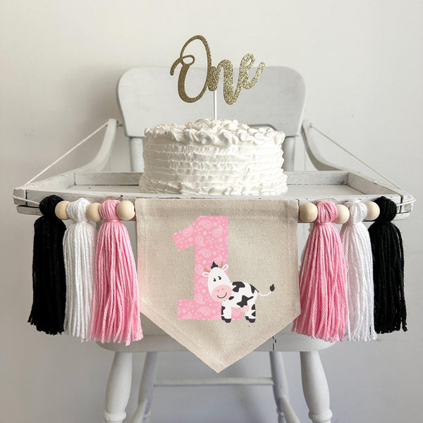 A 5 by 7 inch canvas center flag with a large pink paisley number 1 with a cute black and white cow beside it. On each side there is a pink, white and black 5 inch yarn tassel each separated by a wooden bead for a total of 6 tassels.