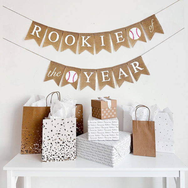 A burlap banner made from 4.5 by 6.5 inch fishtail shaped flags. The top like says ROOKIE in white followed by a baseball image and the word OF on a flag. The bottom line starts with THE on one flag , then a baseball then the word YEAR all in white.