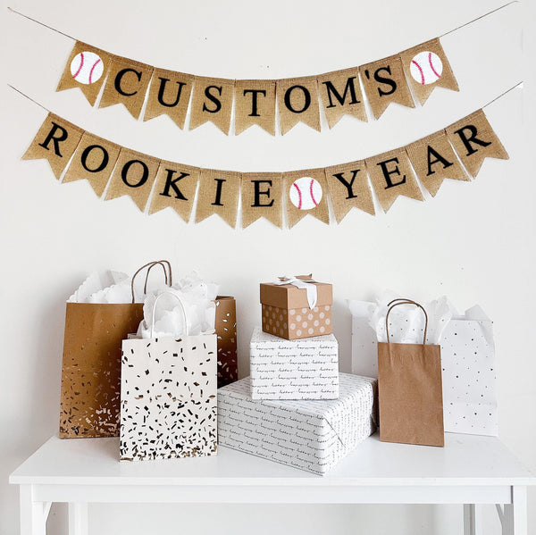 A burlap banner made from 4.5 by 6.5 inch fishtail shaped flags. The top line can be customized with a name in black letters and has a baseball image on each end. The bottom line says ROOKIE YEAR in black letters and has a baseball in the middle.