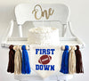 A 5 by 7 inch canvas center flag with a football and the words FIRST above it and the word DOWN below it in royal blue. There is a royal blue, tan and brown 5 inch yarn tassel on each side, each separated by a wooden bead for a total of 6 tassels.