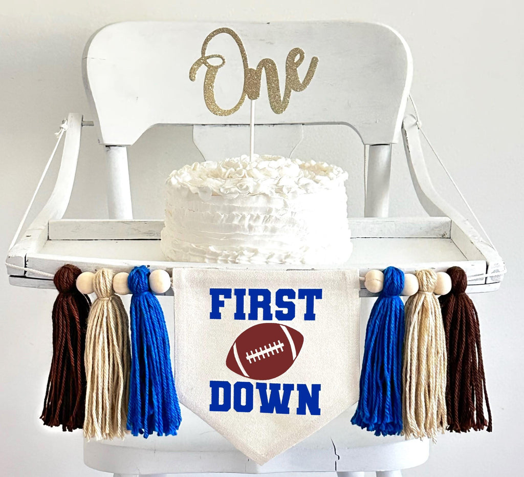A 5 by 7 inch canvas center flag with a football and the words FIRST above it and the word DOWN below it in royal blue. There is a royal blue, tan and brown 5 inch yarn tassel on each side, each separated by a wooden bead for a total of 6 tassels.