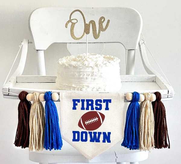 A 5 by 7 inch canvas center flag with a football and the words FIRST above it and the word DOWN below it in royal blue. There is a royal blue, tan and brown 5 inch yarn tassel on each side, each separated by a wooden bead for a total of 6 tassels.