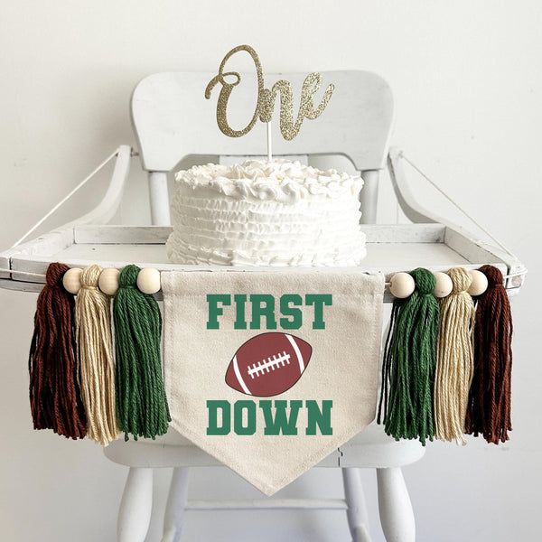 A 5 by 7 inch canvas center flag with the word FIRST in green, a football in the middle, and the word DOWN under the football. On each side there is a dark green, tan and brown 5 inch yarn tassel each separated by a wooden bead for a total of 6.