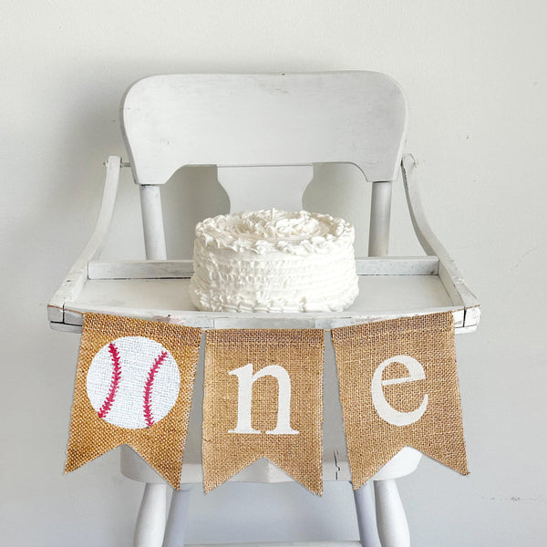 A burlap banner made from three 5 by 7 inch fishtail shaped flags. The first flag has a baseball image on it. The 2nd flag has a white lowercase N, the 3rd flag has a white lowercase letter E. The banner is hung on a high chair in the photo.