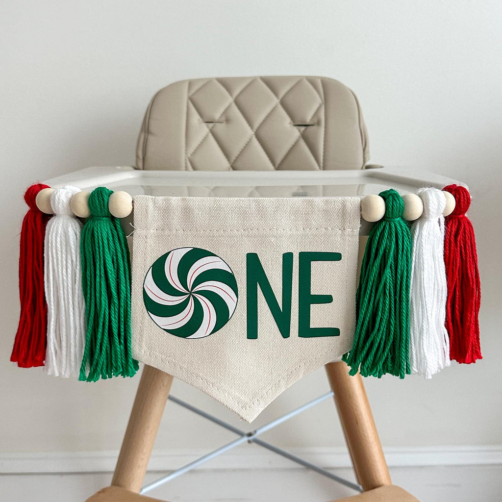 A 5 by 7 inch canvas center flag with a green swirl peppermint candy followed by the letters NE in green to spell one. There is a green, white and red 5 inch yarn tassel on each side, each separated by a wooden bead for a total of 6 tassels.