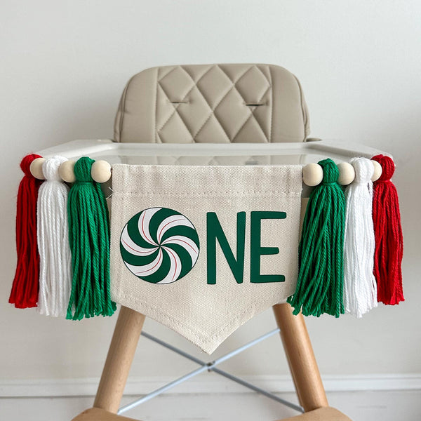 A 5 by 7 inch canvas center flag with a green swirl peppermint candy followed by the letters NE in green to spell one. There is a green, white and red 5 inch yarn tassel on each side, each separated by a wooden bead for a total of 6 tassels.