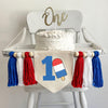 A 5 by 7 inch canvas center flag with a big royal blue number 1 and a red white and blue bomb popsicle propped against it. There is a red, white and blue 5 inch yarn tassel on either side, each separated by a wooden bead.