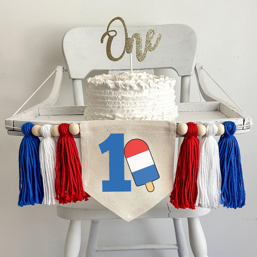 A 5 by 7 inch canvas center flag with a big royal blue number 1 and a red white and blue bomb popsicle propped against it. There is a red, white and blue 5 inch yarn tassel on either side, each separated by a wooden bead.