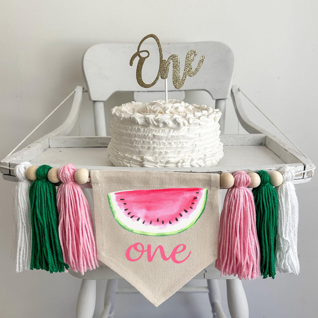 A 5 by 7 inch canvas center flag with a slice of pink watermelon and the word ONE in pink script below it. There are pink, green and white 5 inch yarn tassels on either side each separated by a wooden bead for a total of 6 beads.