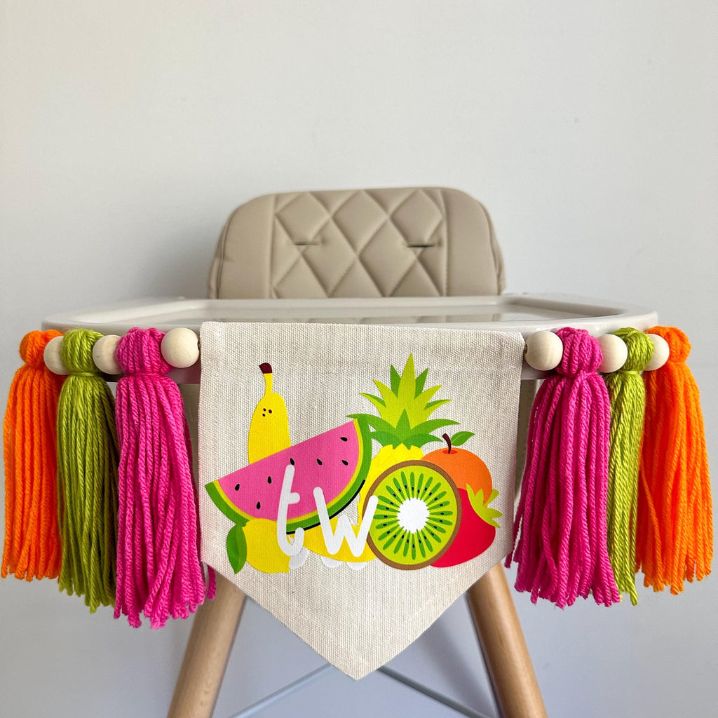 A 5 by 7 inch canvas center flag with a bunch of tropical fruit and the word TWO. There is a 5 inch hot pink, lime green and orange tarn tassel on either side, each separated by a wooden bead for a total of 6 tassels. It is hung on a high chair.