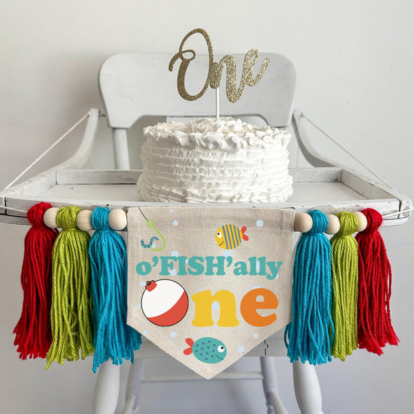 A 5 by 7 inch canvas center flag with the words OFISHALLY ONE with a bobber as the O and 2 tropical fish and bubbles. There is a 5 inch aqua, lime and red colored yarn tassel on each side each separated by a wooden bead for a total of 6 tassels.