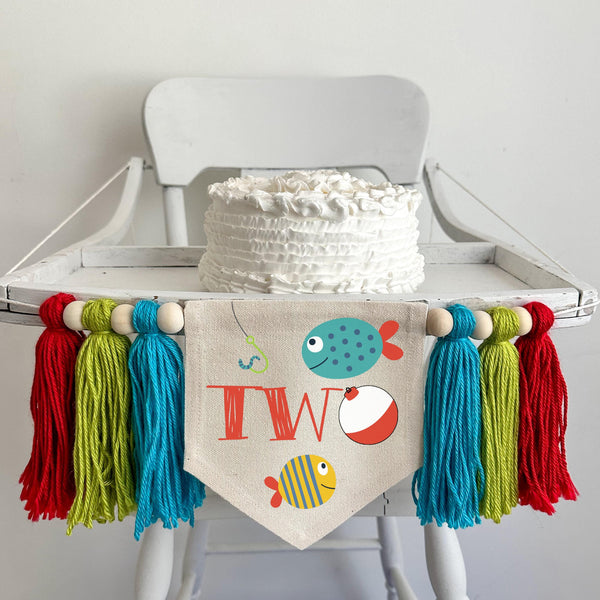 A 5 by 7 inch canvas center flag with the word TWO in red and a bobber as the O. There are 2 tropical fish and a hook. There is a 5 inch aqua, lime and red colored yarn tassel on each side each separated by a wooden bead for a total of 6 tassels.