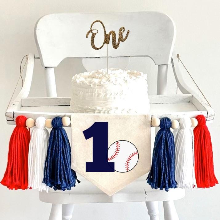 A 5 by 7 inch canvas banner with a large navy blue number 1 with a baseball beside it. There is a 5 inch navy blue, white and red yarn tassel on each side with a wooden bead between each of them for a total of 6 tassels.
