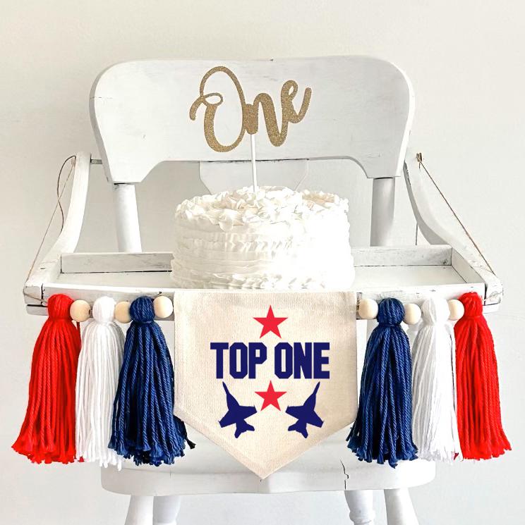 A 5 by 7 inch canvas banner with the words TOP ONE in navy with red stars and navy planes. There is a 5 inch navy blue, white and red yarn tassel on each side with a wooden bead between each of them for a total of 6 tassels.