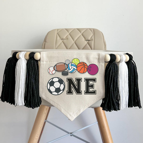 A 5 by 7 inch canvas center flag with a soccer ball and the letters NE in black with a row of sports balls at the top. There is a 5 inch black, white and black colored yarn tassel on each side each separated by a wooden bead for a total of 6 tassels.