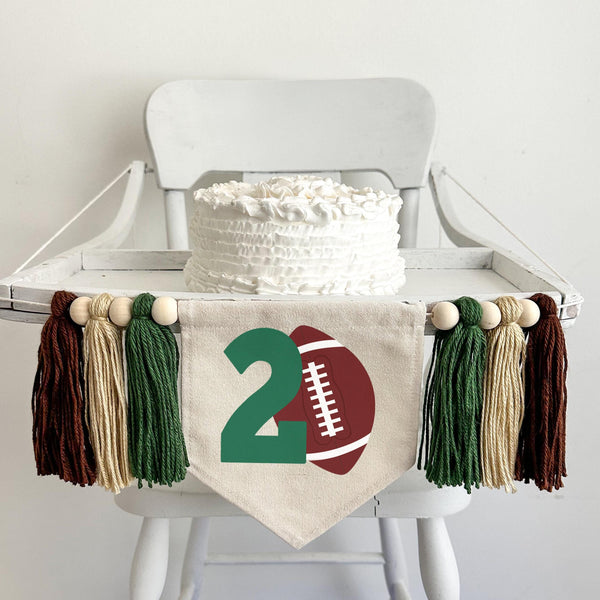 A 5 by 7 inch canvas center flag with a football and a large green number 2. There is a 5 inch dark green, tan and brown colored yarn tassel on each side each separated by a wooden bead for a total of 6 tassels. It is displayed on a high chair.