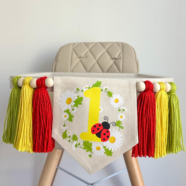 A 5 by 7 inch canvas center flag with a yellow number 1 and a lady bug, surrounded by a daisy wreath. There are 5 inch red, yellow and lime green yarn tassels on each side for a total of 6 tassels. The banner is shown on a high chair.