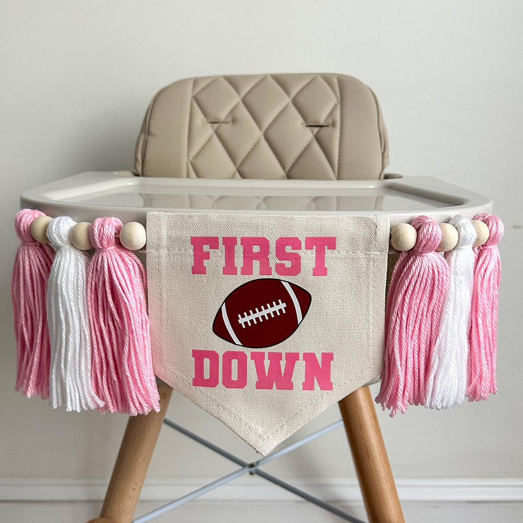 A 5 by 7 inch canvas banner with the words FIRST DOWN in pink with an image of a football between the words. There is a 5 inch pink, white and pink yarn tassel on each side with a wooden bead between each of them for a total of 6 tassels.